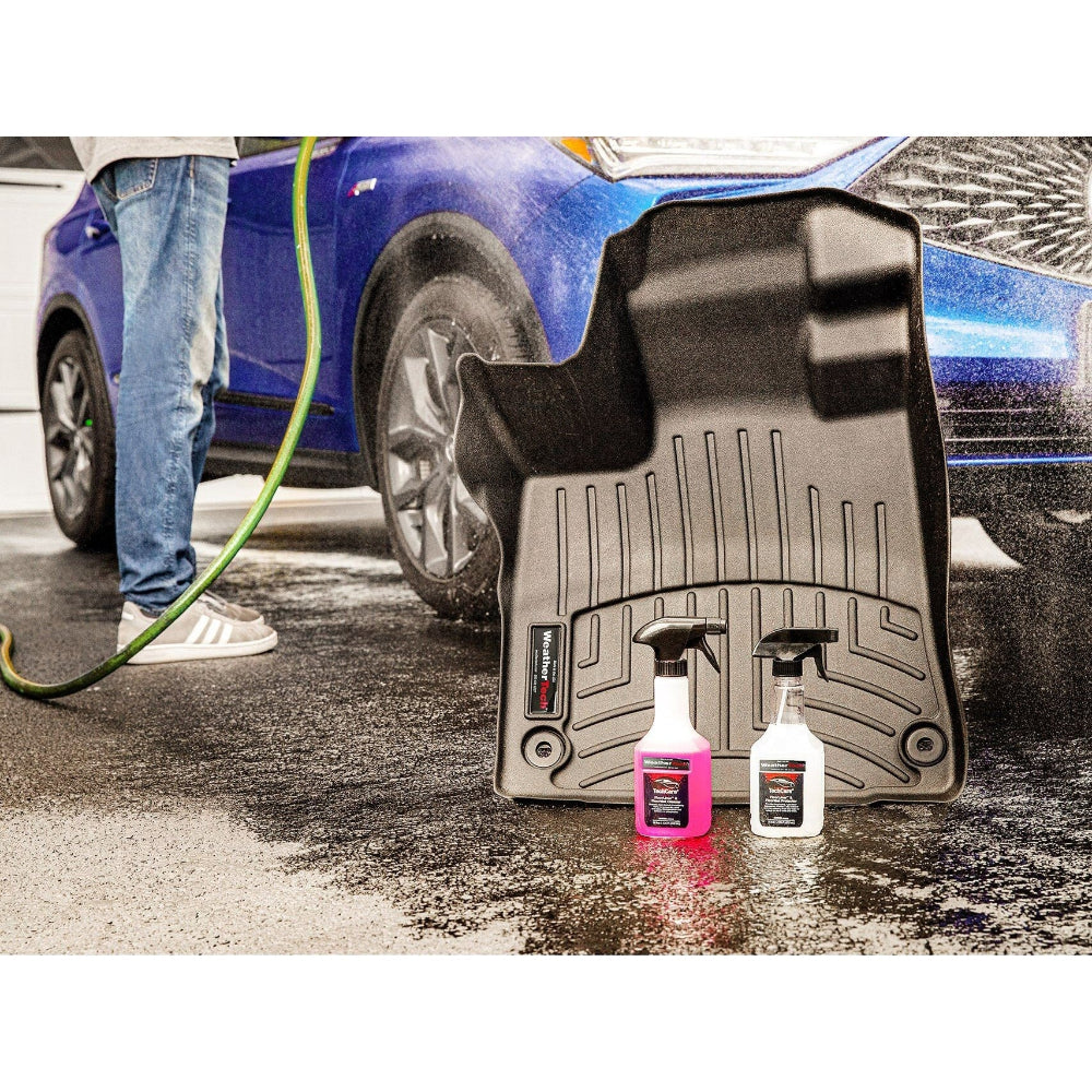 Person cleaning a car with a hose, showing a set of car mats and cleaning products on the ground.