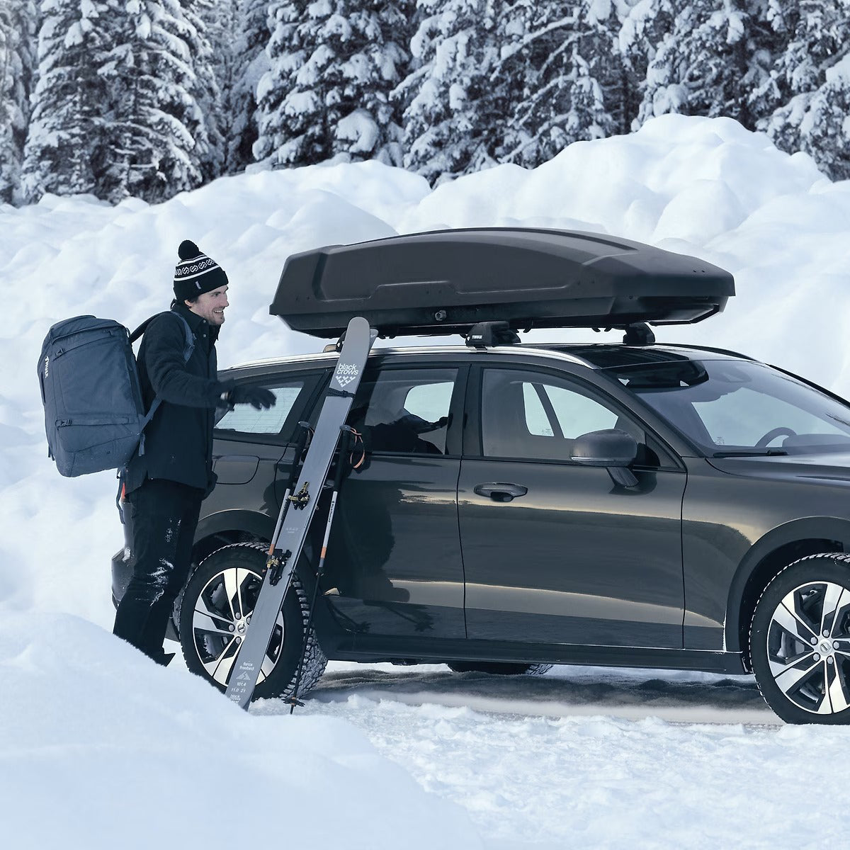 Person with a backpack loading a car with a roof box in a snowy landscape