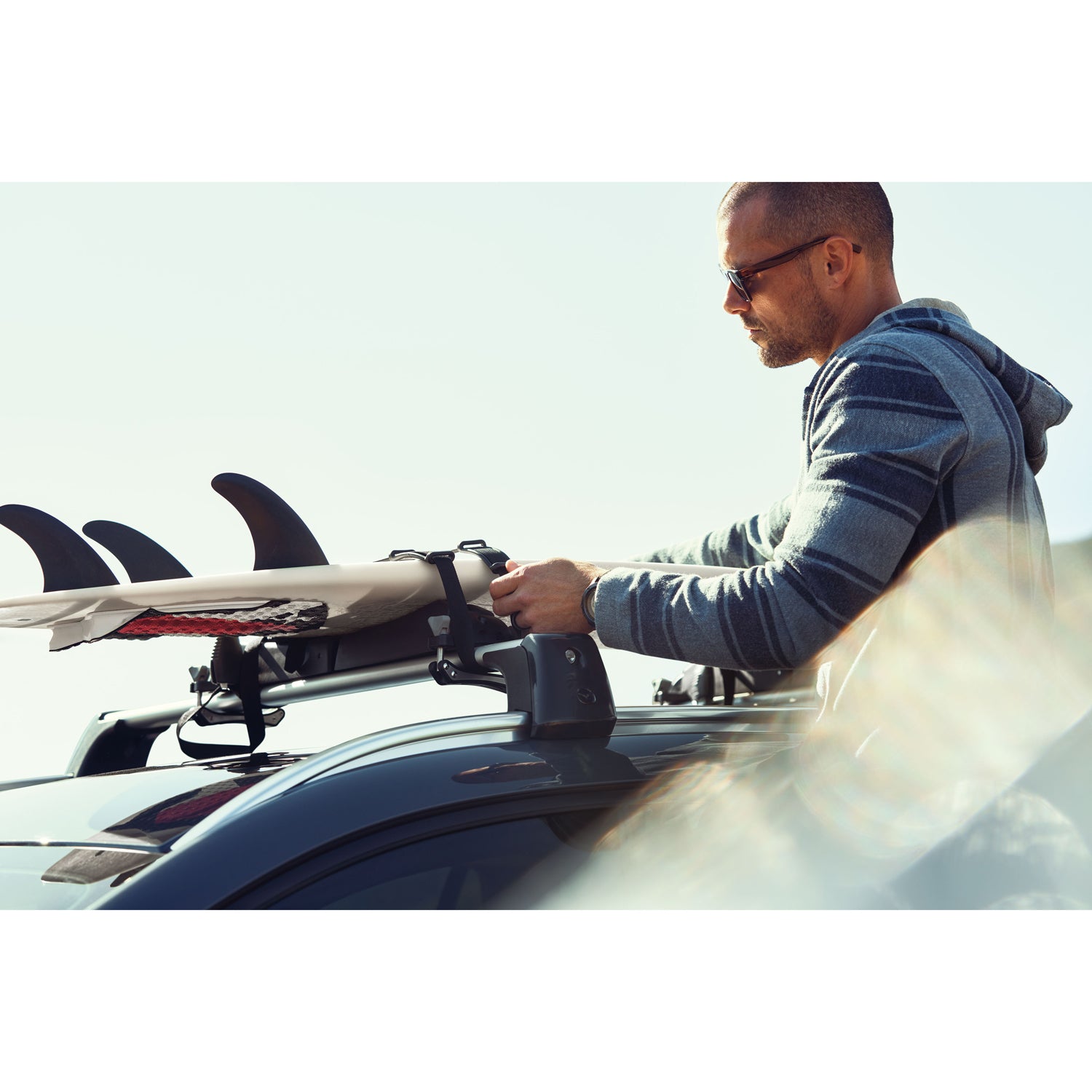 Man securing a surfboard to a car roof rack with a clear sky background